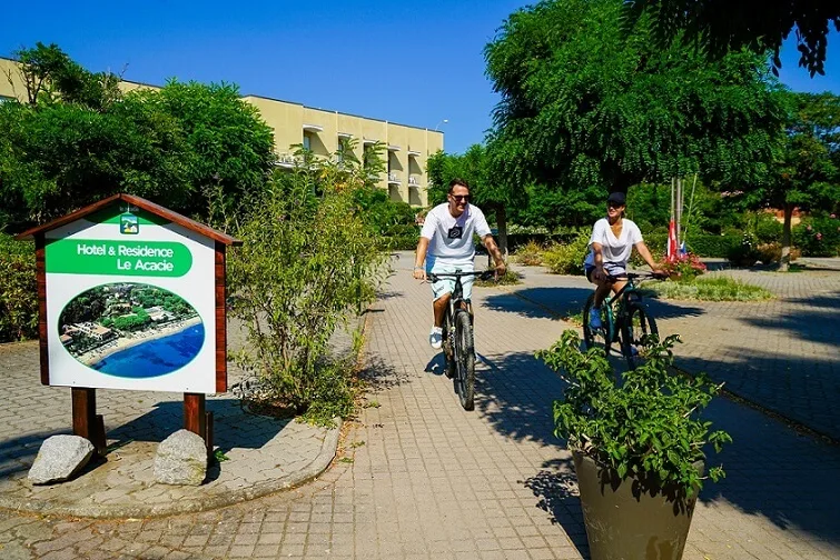 Coppia in bicicletta sul vialetto dell'hotel all'Isola d'Elba, tra piante verdi e cartello informativo